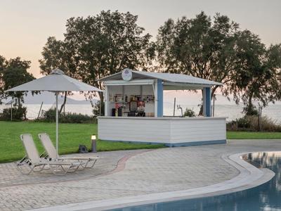 Area piscina all'aperto con lettini bianchi, ombrellone e bar a bordo piscina con alberi sullo sfondo.