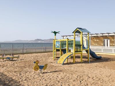 Parco giochi sulla spiaggia con scivolo e struttura per arrampicarsi vicino al mare e area recintata.