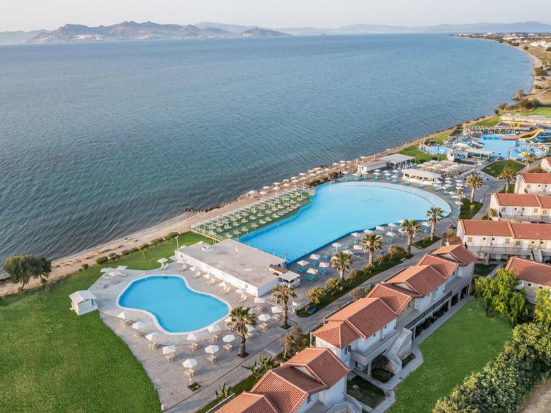 Vista aerea di un resort sul mare con piscine, lettini prendisole e edifici con tetti rossi.