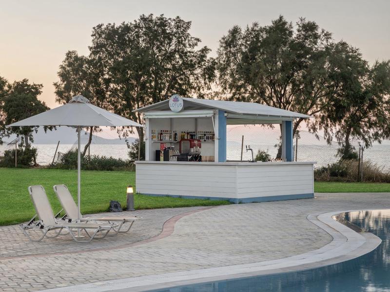 Area piscina all'aperto con lettini bianchi, ombrellone e bar a bordo piscina con alberi sullo sfondo.