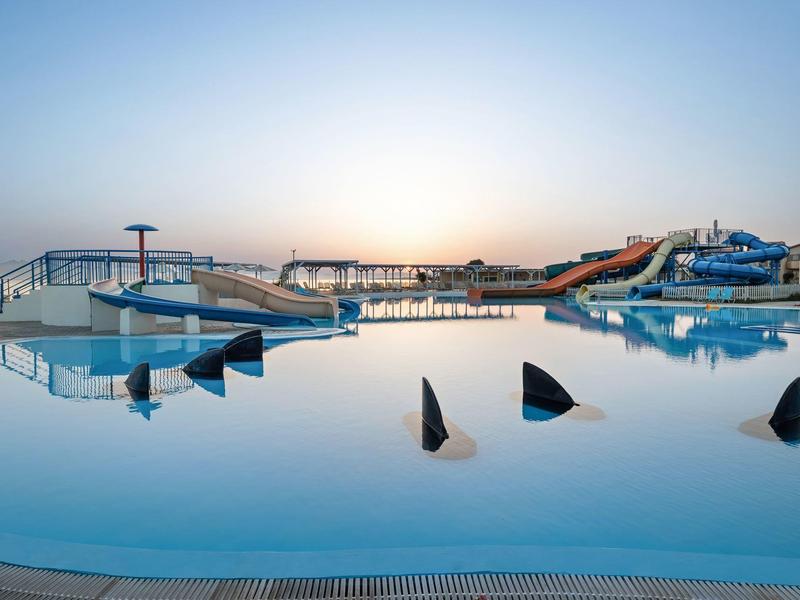 Piscina con decorazioni a pinna di squalo e scivoli d’acqua sullo sfondo sotto un cielo limpido.