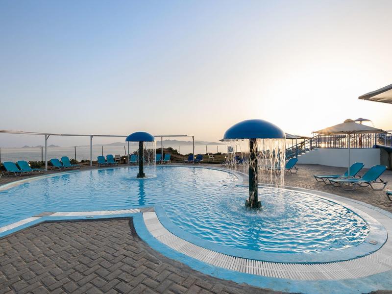 Piscina all'aperto con fontane ad ombrello e sdraio sotto un cielo limpido al tramonto.