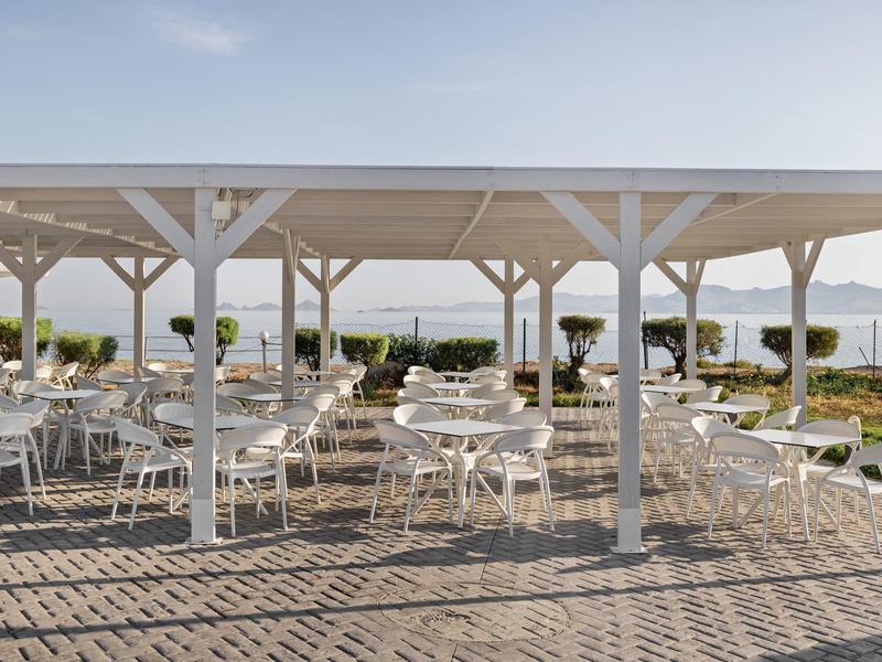 Area pranzo all'aperto con tavoli e sedie bianchi sotto una pergola vicino al mare.