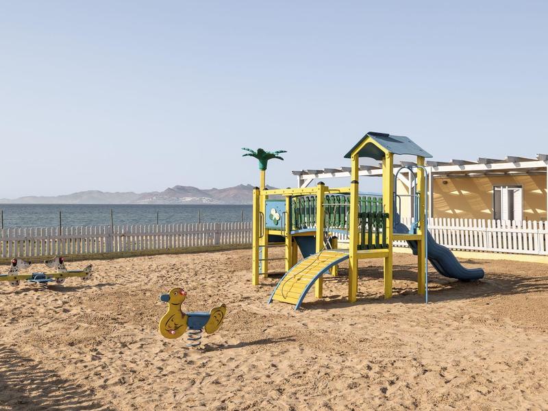 Parco giochi sulla spiaggia con scivolo e struttura per arrampicarsi vicino al mare e area recintata.