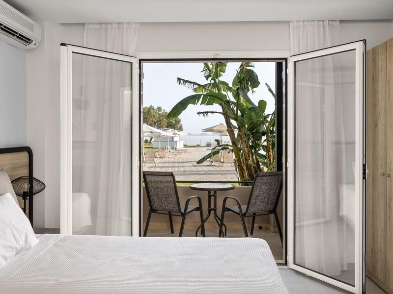 Camera luminosa con apertura su balcone con tavolo, sedie e piante tropicali, vista mare.