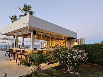 Moderner Außenbereich einer Strandbar mit Sitzgelegenheiten und tropischer Bepflanzung bei Sonnenuntergang.