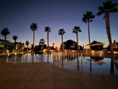Area piscina di un hotel al tramonto con palme e lettini illuminati.