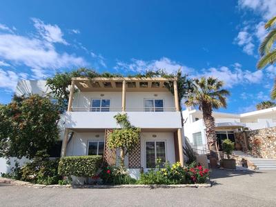 Casa vacanze bianca su due piani con terrazza, palme e piante fiorite sotto un cielo azzurro.
