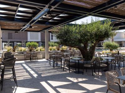 Überdachte Terrasse mit großem Baum in der Mitte, umgeben von Tischen, Stühlen und Pflanzen.