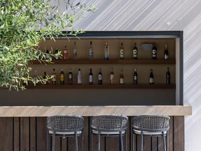 Area bar esterna moderna con bancone in legno, tre sgabelli e scaffale con bottiglie di liquore.