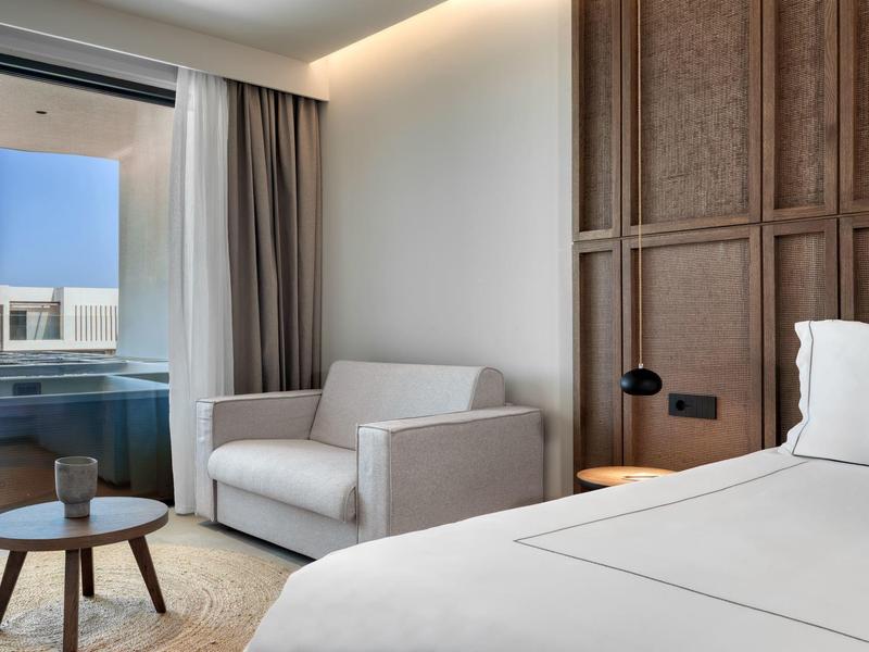 Modern hotel room with bed, sofa, small table, and balcony with sea view.