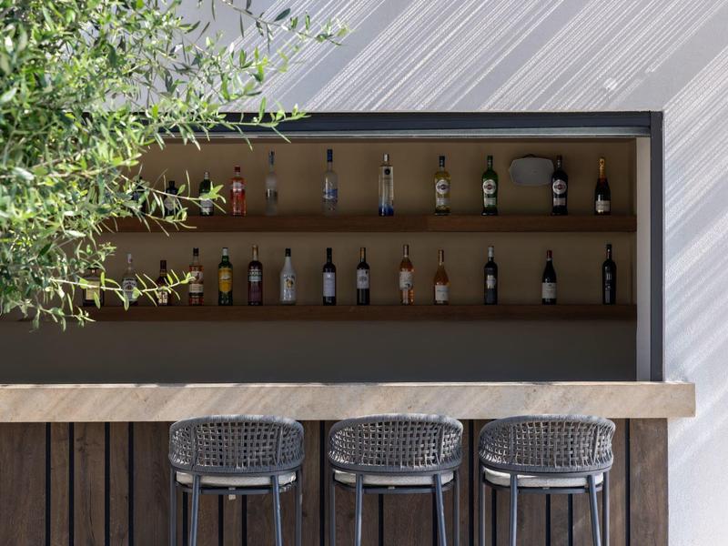 Modern outdoor bar area with wooden counter, three stools, and illuminated bottle shelves.