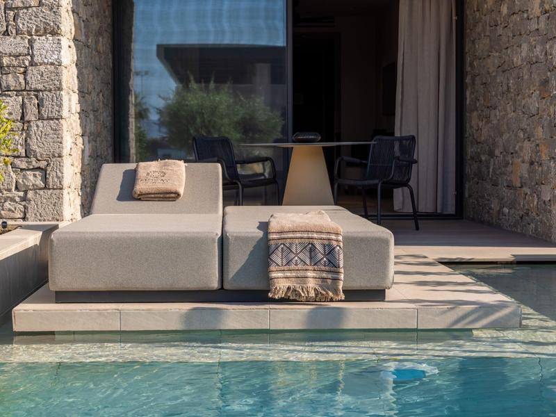Two gray lounge chairs with cushions and a blanket beside a pool in front of a stone house with a terrace.