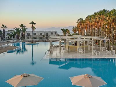 Widok na basen hotelowy z parasolami, palmami i budynkami o zachodzie słońca.