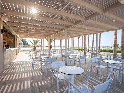 Offener Gastronomiebereich mit weißen Tischen und Stühlen unter einer überdachten Terrasse mit Blick auf Palmen.