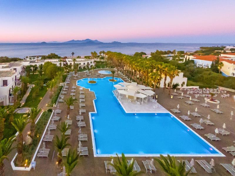 Duży basen na zewnątrz z leżakami i palmami w kompleksie hotelowym z widokiem na morze o zachodzie słońca.