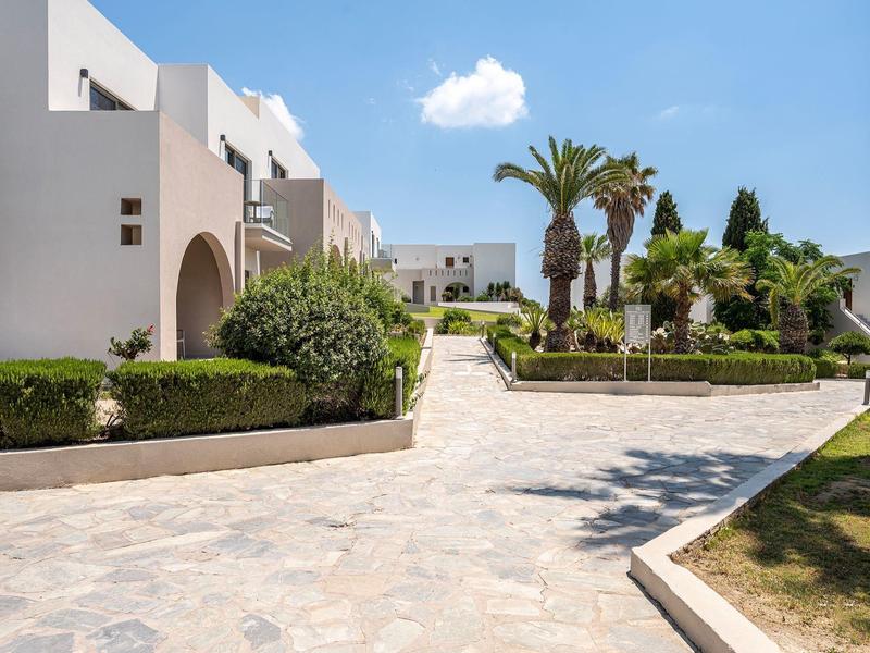 Nowoczesny kompleks hotelowy z białymi budynkami, palmami i brukowanym dziedzińcem pod niebieskim niebem.