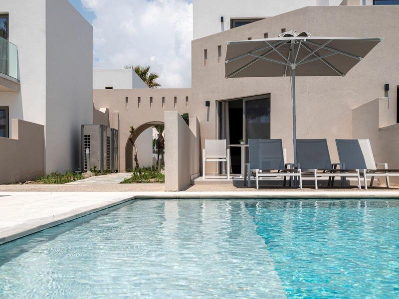 Nowoczesny basen z leżakami i parasolem przed białym budynkiem.