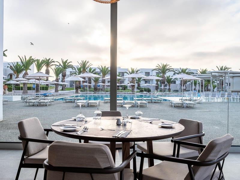 Okrągły stół z czterema krzesłami na balkonie z widokiem na basen i palmy w ośrodku.