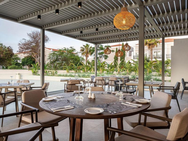 Otwarta restauracja z drewnianymi stołami i krzesłami obok basenu i palm.