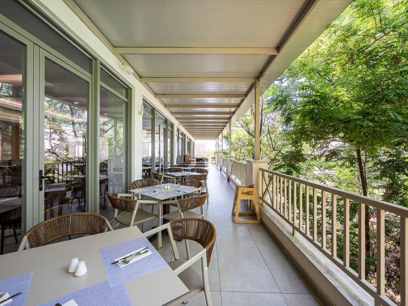 Balkon z stołami i krzesłami obok przeszklonych okien, otoczony zielonymi drzewami.