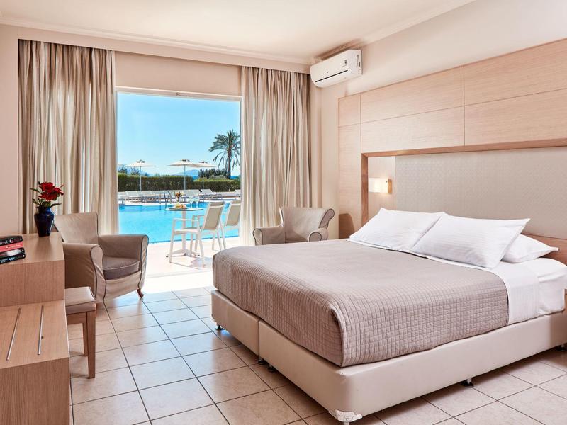 Jasny pokój hotelowy z podwójnym łóżkiem, fotelami i widokiem na basen i palmy