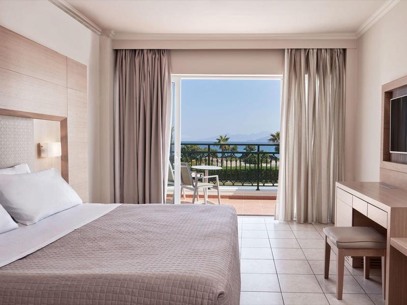 Jasny pokój hotelowy z podwójnym łóżkiem i widokiem na taras i morze.