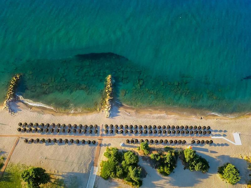 Widok z lotu ptaka na piaszczystą plażę z parasolami i leżakami przy czystej, niebieskiej wodzie.