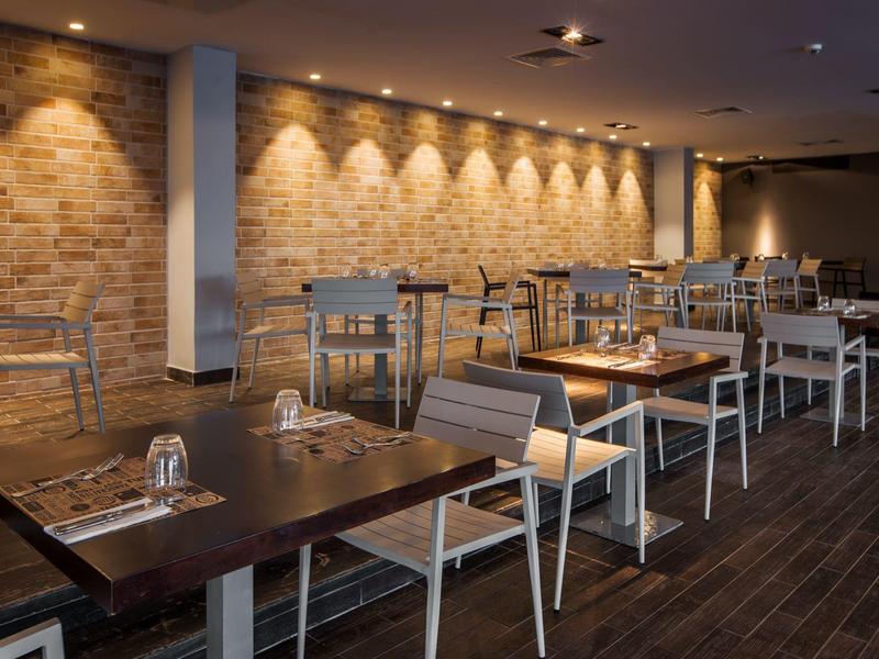 Nowoczesna restauracja z drewnianymi stołami, białymi krzesłami i ciepłym oświetleniem ściennym.