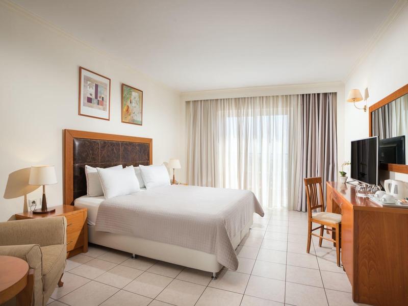 Jasny pokój hotelowy z dużym łóżkiem, sofą, biurkiem i telewizorem obok okna z zasłonami.