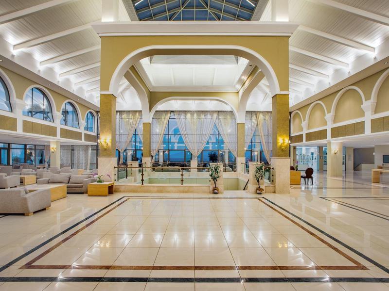 Przestronna lobby hotelowa z białymi siedzeniami, złotymi kolumnami i wysokimi sufitami z podświetlanymi łukami.