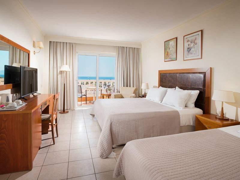 Pokój hotelowy z dwoma łóżkami, telewizorem, biurkiem i balkonem z widokiem na morze.