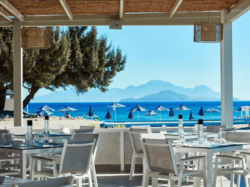 Otwarty taras restauracyjny z widokiem na morze i łańcuch górski w tle.