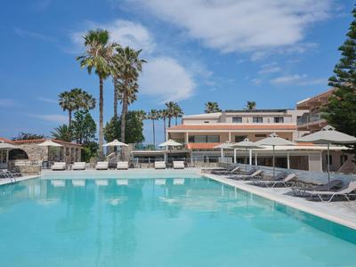 Een groot zwembad met ligstoelen en palmbomen voor een meerlaagse hotelgebouw onder een blauwe lucht.