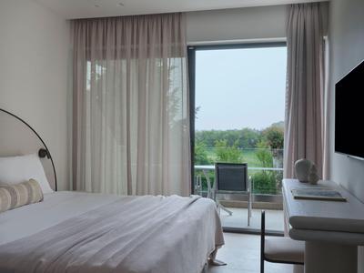 Modernes Hotelzimmer mit Doppelbett, Vorhängen und Blick auf einen Balkon mit Stuhl und grünem Ausblick.