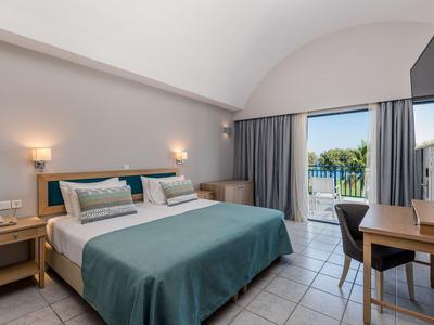 Helles Hotelzimmer mit Doppelbett, Schreibtisch, Stühlen und Blick auf eine Terrasse mit Pflanzen.