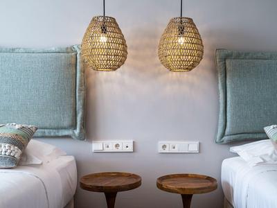 Zwei Einzelbetten mit blauen Polsterkopfteilen und zwei runden Holztischen, darüber hängen zwei geflochtene Lampen.