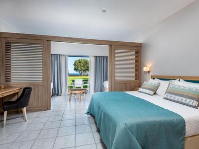 Helles Hotelzimmer mit großzügigem Bett, Schreibtisch und Blick auf Terrasse und Meer.