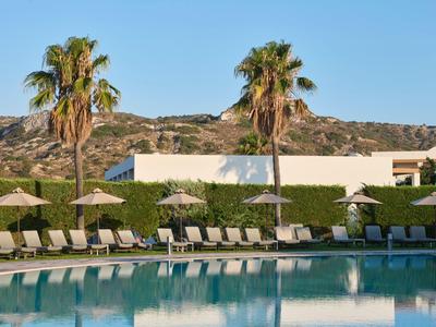 Pool mit Liegestühlen, Strohhüten und Palmen vor Gebäuden und Bergen bei klarem Himmel.