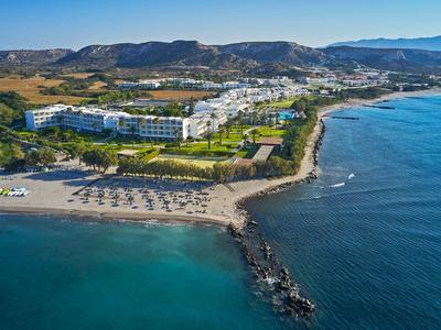 Große Hotelanlage an der Küste mit Strand, Liegestühlen und Berglandschaft im Hintergrund.