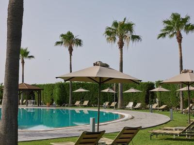 Runder Hotelpool mit Liegestühlen und Sonnenschirmen, umgeben von Palmen.