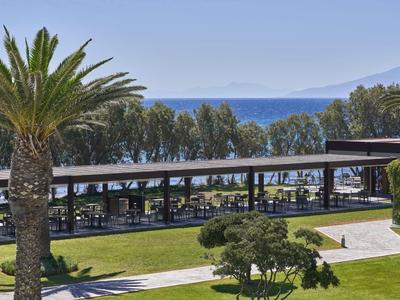 Blick auf einen Hotelgarten mit Palmen, überdachtem Sitzbereich und Meer im Hintergrund.