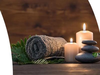 Setup benessere con candele accese, asciugamano grigio e pietre nere su superficie di legno.