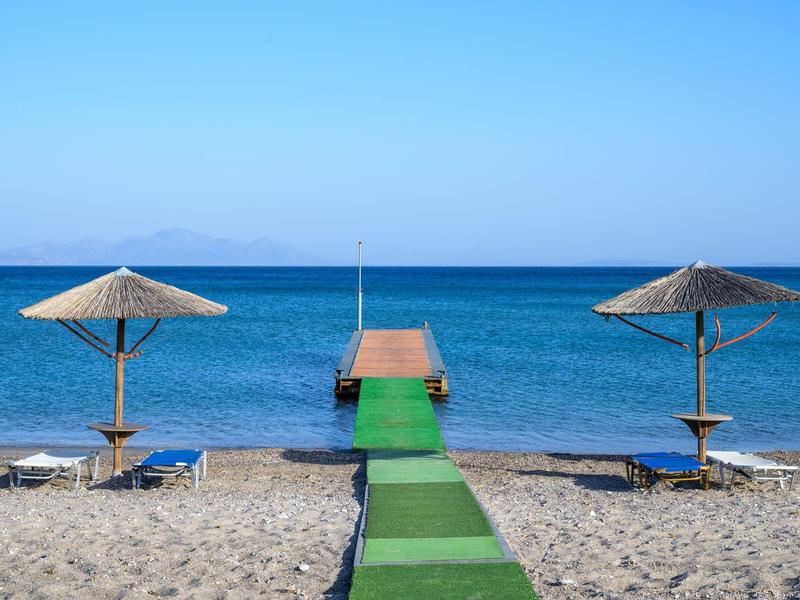 Spiaggia sabbiosa con due ombrelloni di paglia e un molo verde che si estende nel mare blu.