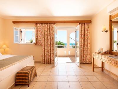 Chambre d'hôtel lumineuse avec lit, banc, rideaux et accès au balcon avec vue mer.