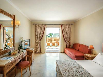 Coin de chambre d'hôtel lumineux avec lit double, canapé, bureau en bois et balcon avec vue extérieure.