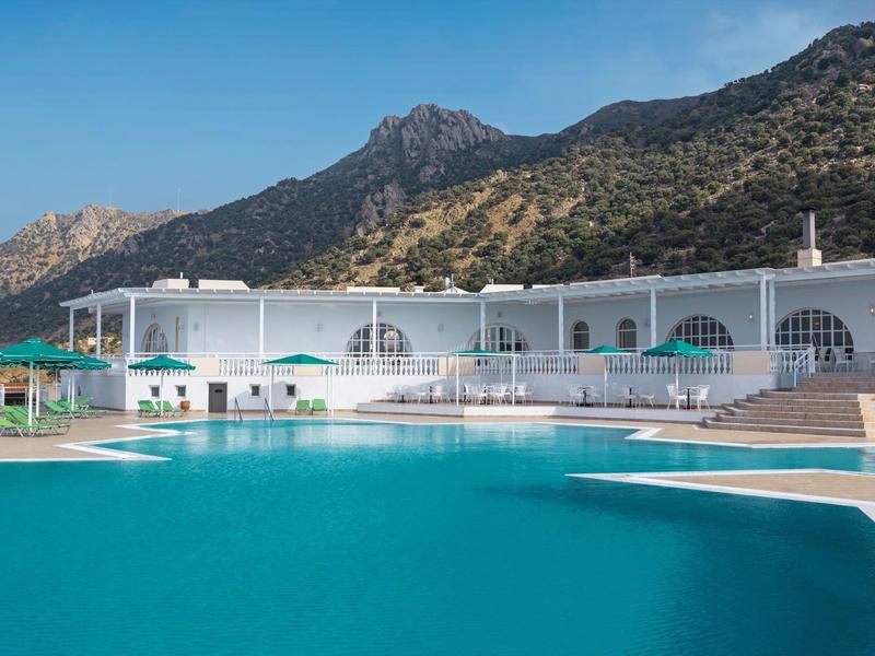 Piscine d'hôtel moderne avec bâtiments blancs, montagnes en arrière-plan et ciel bleu clair.