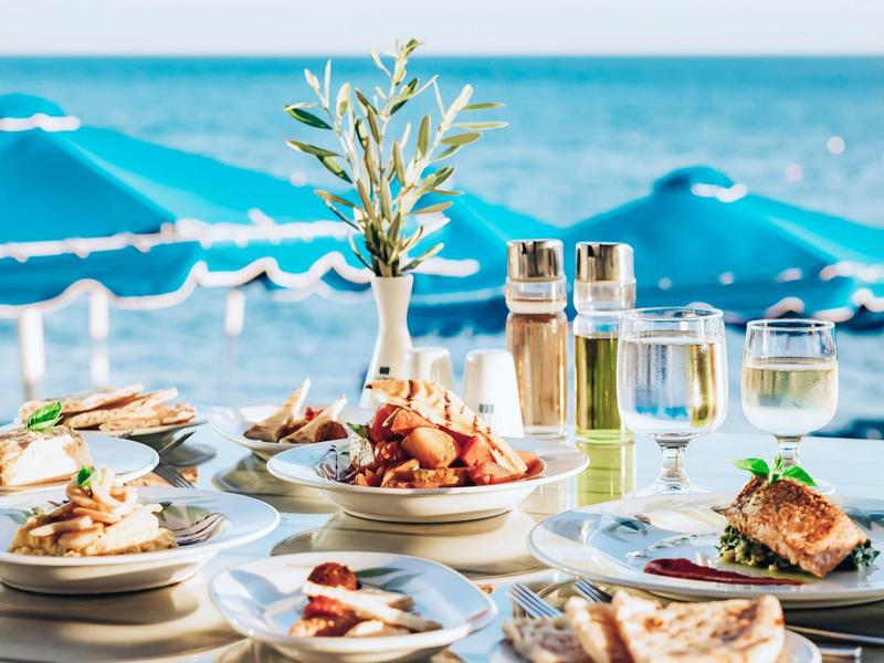 Table avec divers plats et boissons au bord de la mer, parasols bleus en arrière-plan.