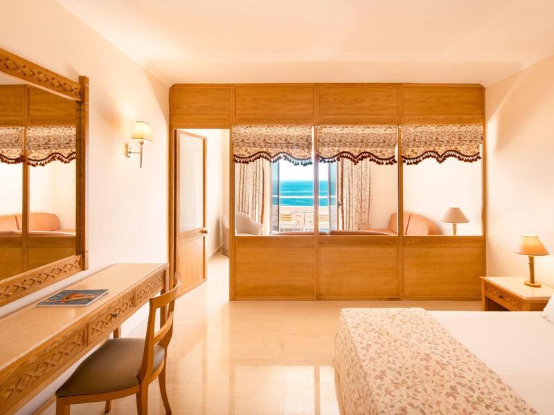 Chambre d'hôtel lumineuse avec lit, miroir, bureau et vue sur la mer à travers de grandes fenêtres.