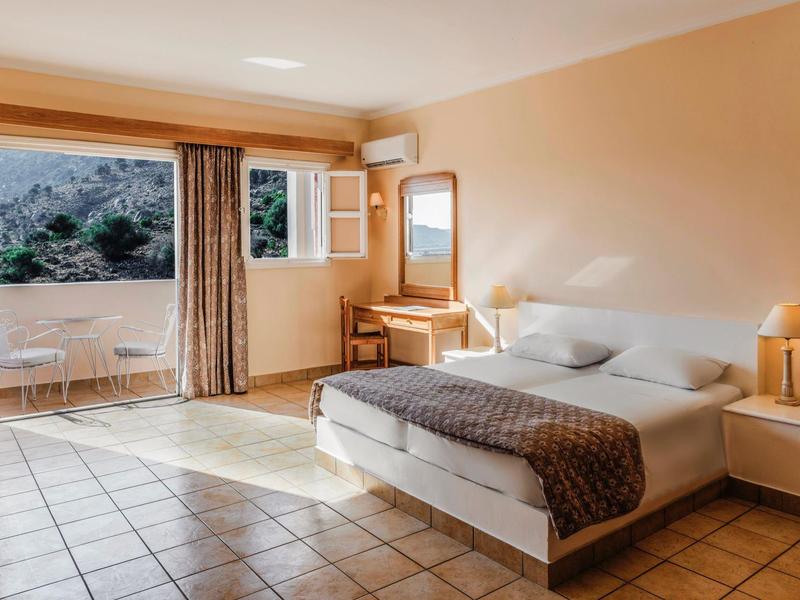 Chambre d'hôtel lumineuse avec lit double, bureau, tables de chevet et terrasse avec vue sur la montagne.